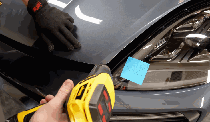 A technician uses a heat gun to apply paint protection film to the edge of a vehicle hood, while another person presses down the film with a gloved hand.
