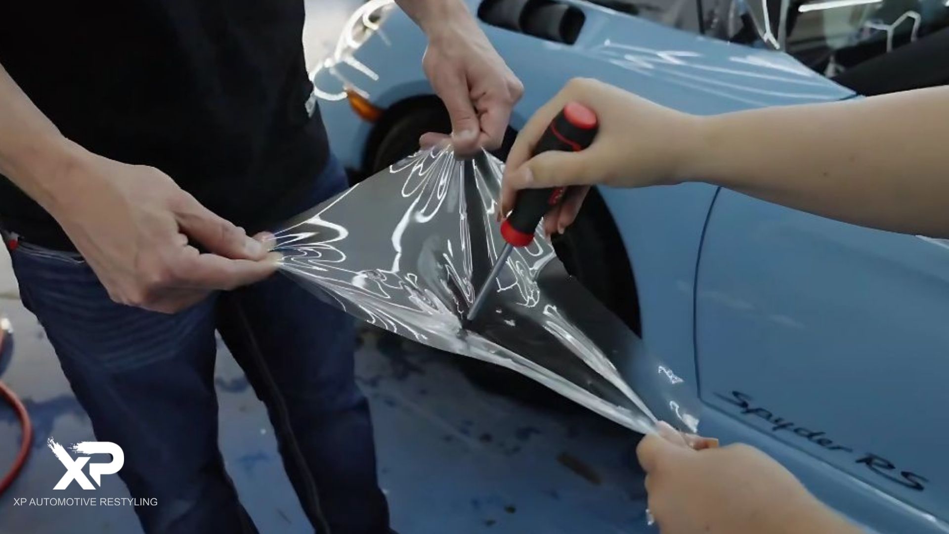 Two people stretch a clear sheet of paint protection film while testing its durability by puncturing it with a screwdriver, with a blue Porsche Spyder RS in the background.