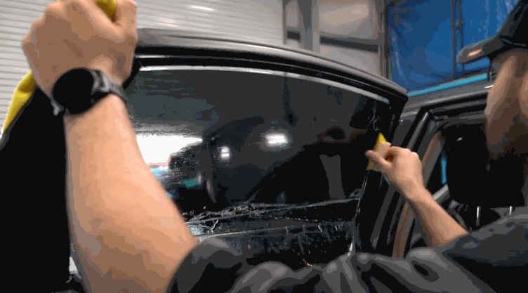 A person presses and smooths window tint film onto a large car door window using a yellow tool, with water droplets visible behind the film.