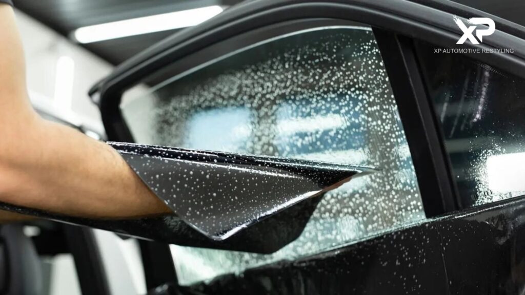 A partially installed tint film being laid over a wet car window, with water droplets covering the glass for smooth application.