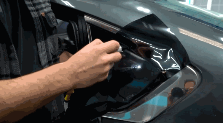 A close-up of a technician applying dark window tint film to a car's rear quarter window using a squeegee tool to smooth it out.