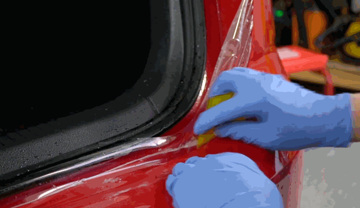 Close-up of gloved hands applying Paint Protection Film to the edge of a red vehicle near the trunk using a yellow squeegee. The film is being smoothed to eliminate air bubbles and ensure clean adhesion.