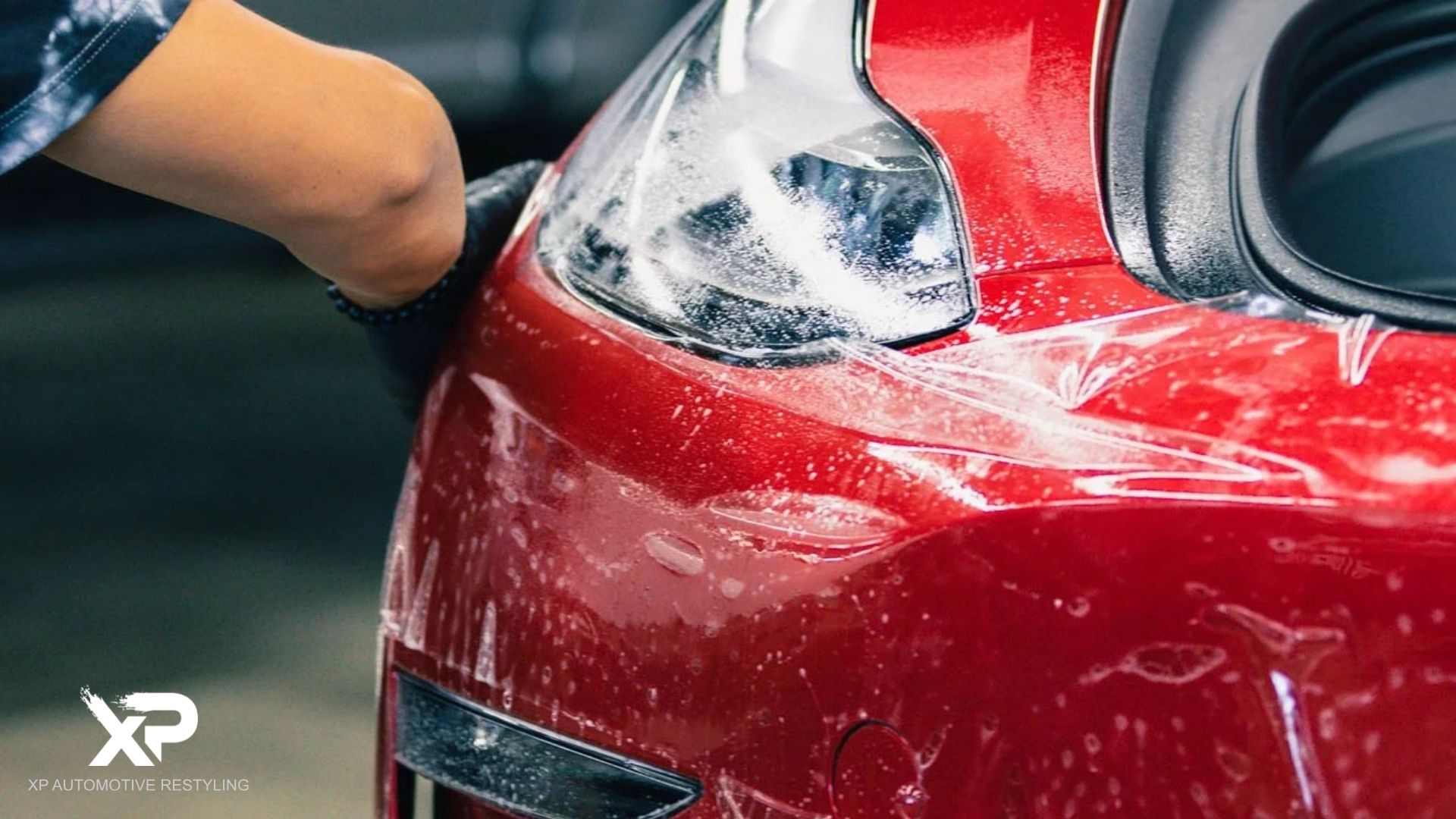 Paint Protection Film being applied to the headlight and front fender of a red car, with a wet application method visible through the moisture and bubbles on the surface.