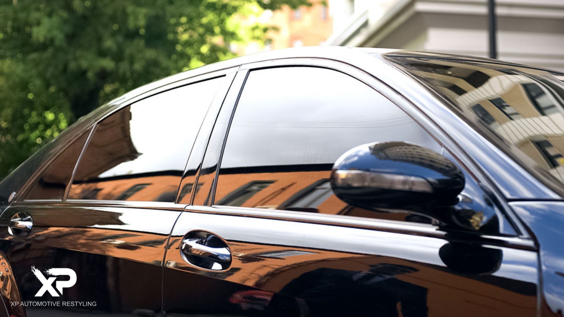 A sleek black sedan with professionally installed dark window tint reflects city buildings and trees on its polished surface.