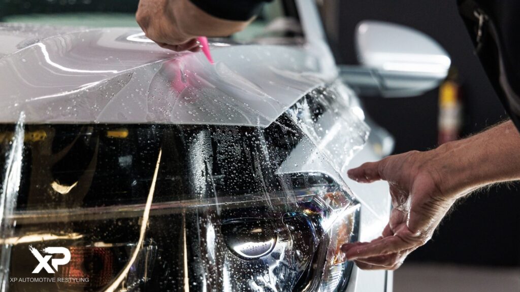 Close-up of a technician applying a paint protection film to the front of a white vehicle, carefully smoothing the transparent film over the headlight and hood to prevent scratches and damage.