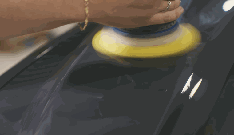 Hand holding a dual-action polisher with a yellow pad buffing a glossy car hood as part of the ceramic coating prep process.