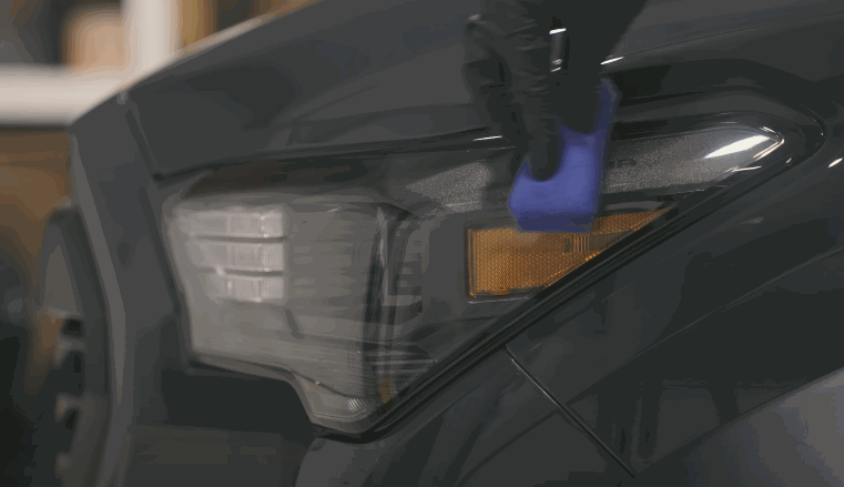 Close-up of a person in black gloves applying ceramic coating to a vehicle’s headlight using a blue applicator pad.