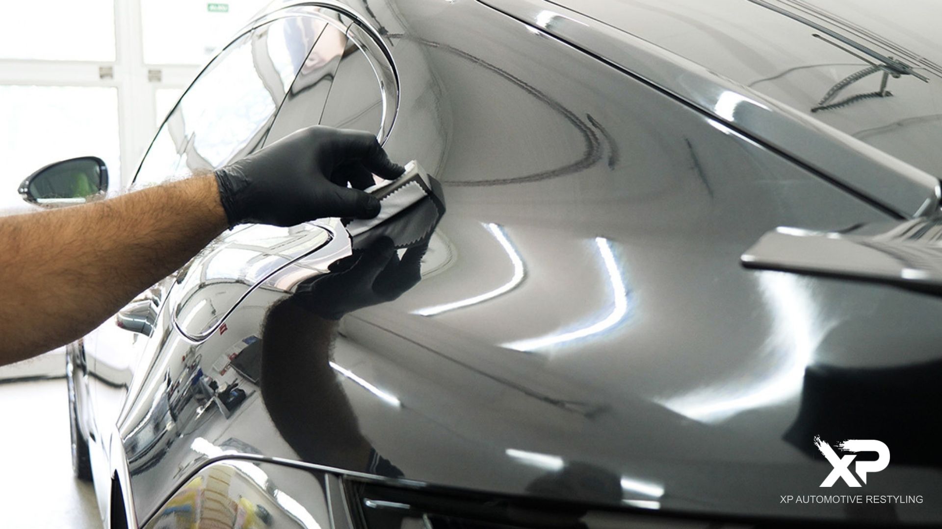 Technician wearing black gloves applying ceramic coating to the rear quarter panel of a sleek black car using an applicator block.