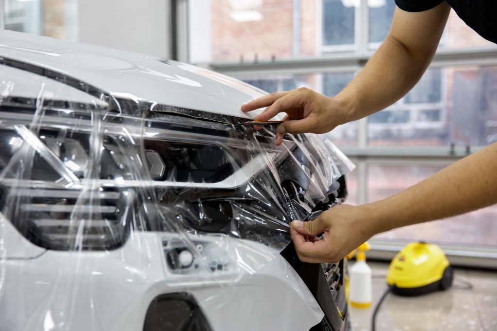 Technician applying clear paint protection film to the front bumper and headlight of a white car during the PPF installation process.