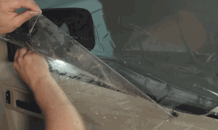 Close up of hands trimming window tint film along the edge of a car window using a small yellow tool for a precise fit. The glass surface is wet to help position the window tint during installation.