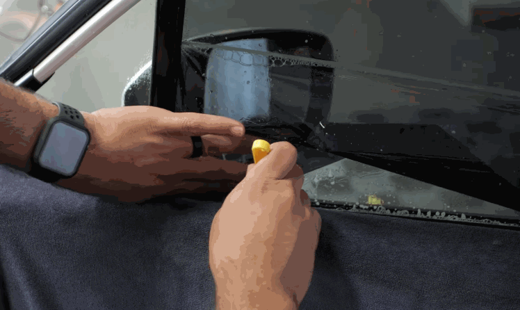 Close up of hands trimming window tint film along the edge of a car window using a small yellow tool for a precise fit. The glass surface is wet to help position the window tint during installation.