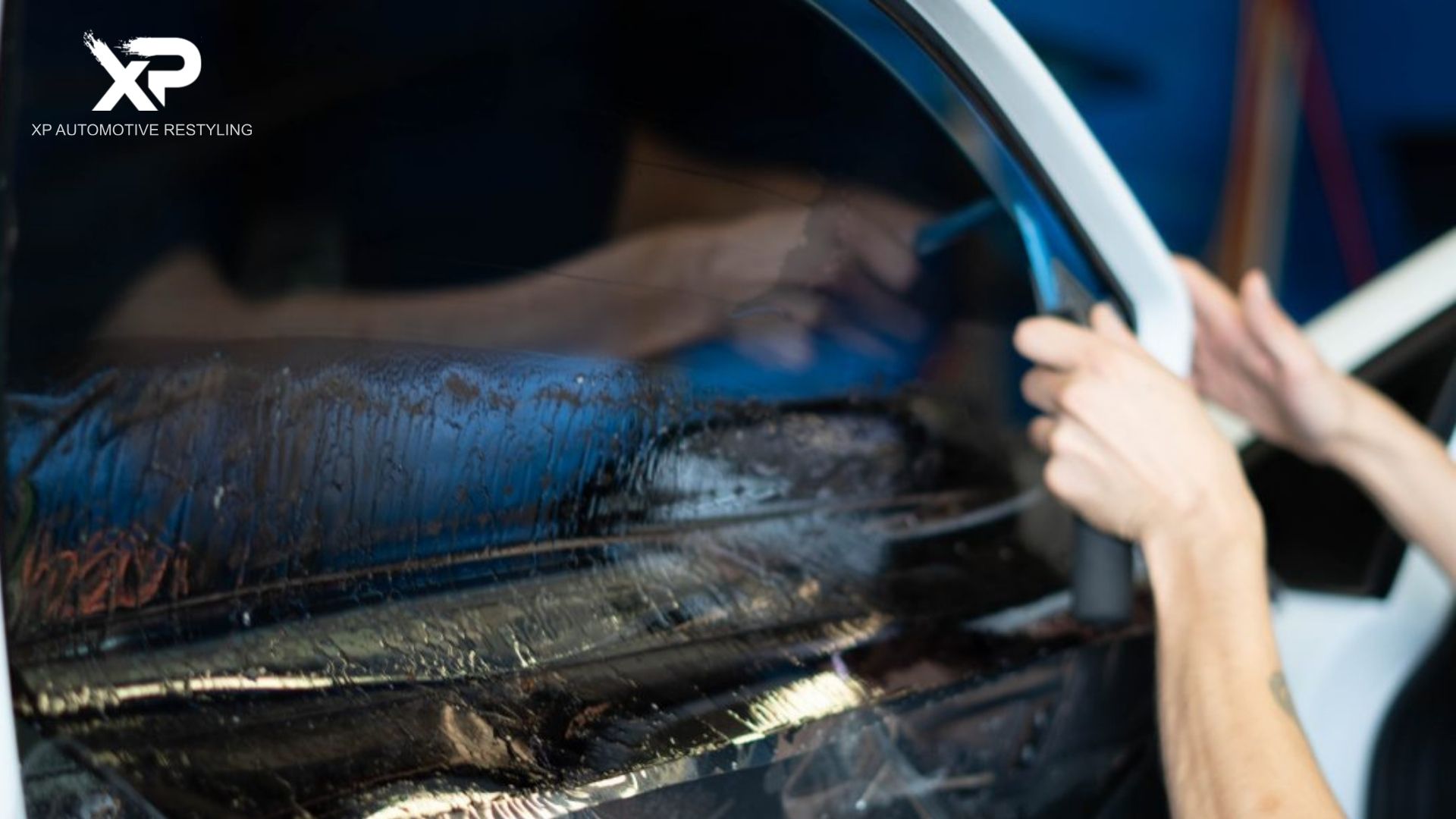 Technician uses a squeegee to smooth window tint across a car door window with visible water and streaks during installation. The top left shows the text "XP Automotive Restyling" branding the window tint service.