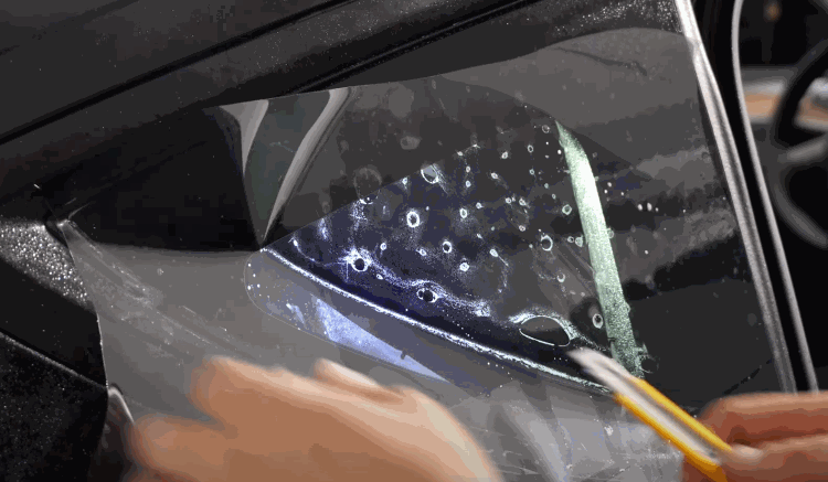 Close up of window tint film being applied to a car window with water droplets and a tool smoothing out bubbles during window tint installation.