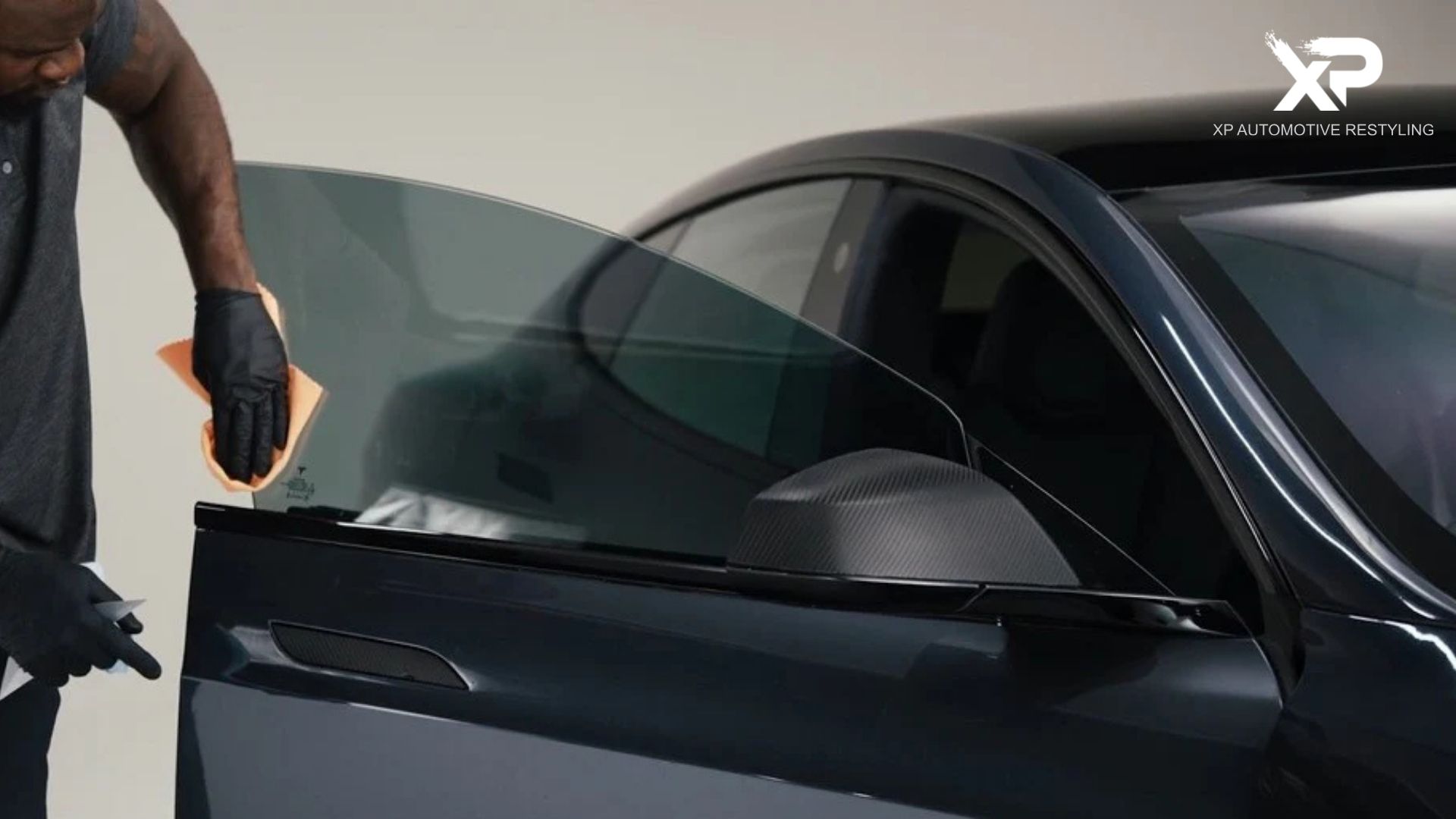 Technician applies window tint film to a car door window using a cloth to smooth the surface for a clean window tint installation with a visible "XP Automotive Restyling" logo.
