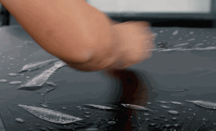 A technician smooths wet paint protection film across the hood of a dark vehicle. Water droplets and film edges show the protective layer being positioned.
