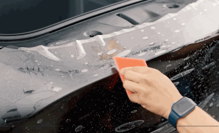 A hand uses an orange squeegee to apply paint protection film along the rear panel of a dark vehicle. Soap bubbles and water help position the clear protective layer.