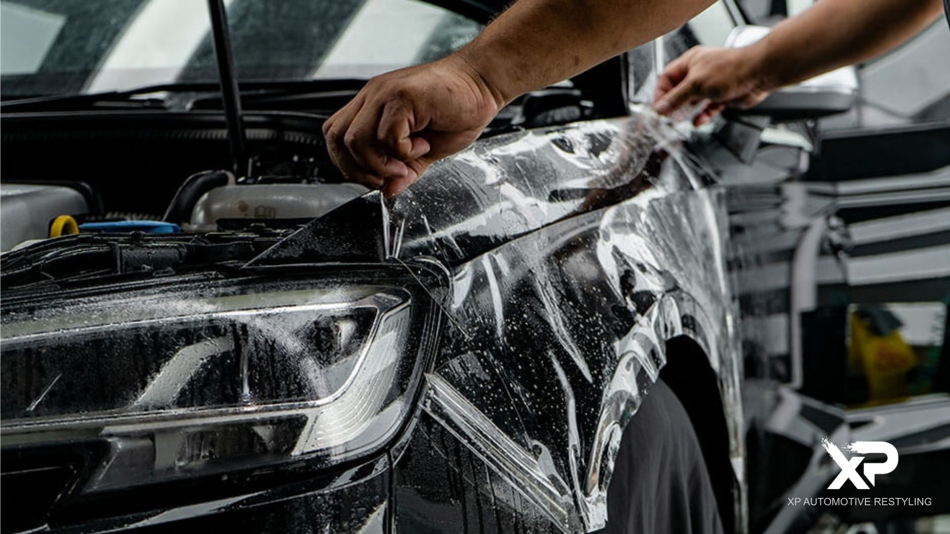 Technicians stretch paint protection film over the front fender and headlight of a black vehicle. The text “XP AUTOMOTIVE RESTYLING” appears in the bottom right corner.