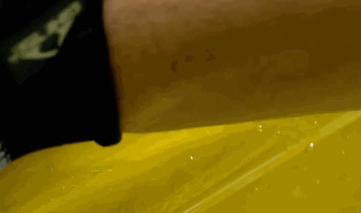 A heat gun warms clear paint protection film on a yellow vehicle panel. The close up shows the film conforming to the paint surface during installation.