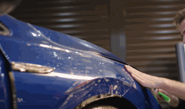 A technician smooths paint protection film over the front fender of a blue vehicle. The clear film is being stretched around the wheel arch to protect the painted panel.