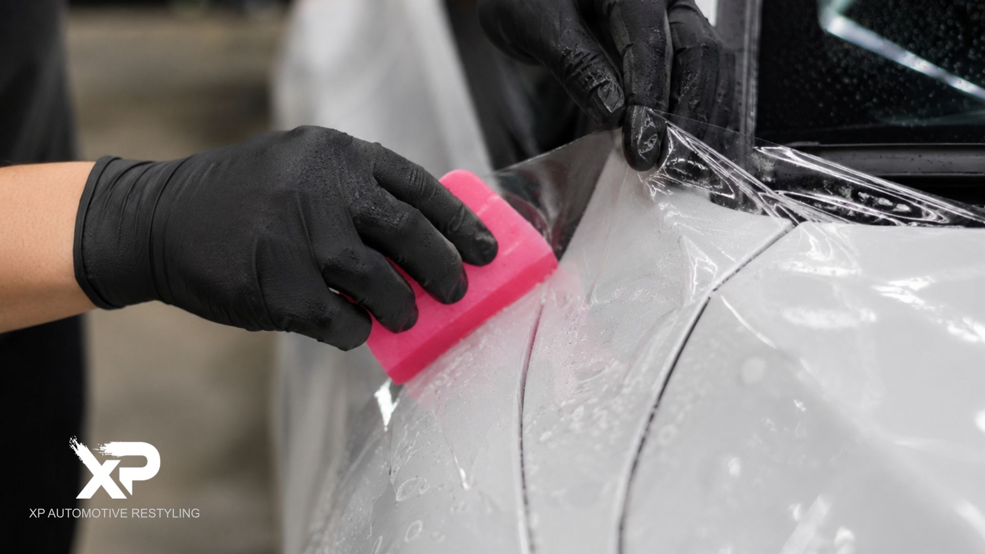 A technician in black gloves uses a pink squeegee to apply paint protection film around the rear panel of a white vehicle.