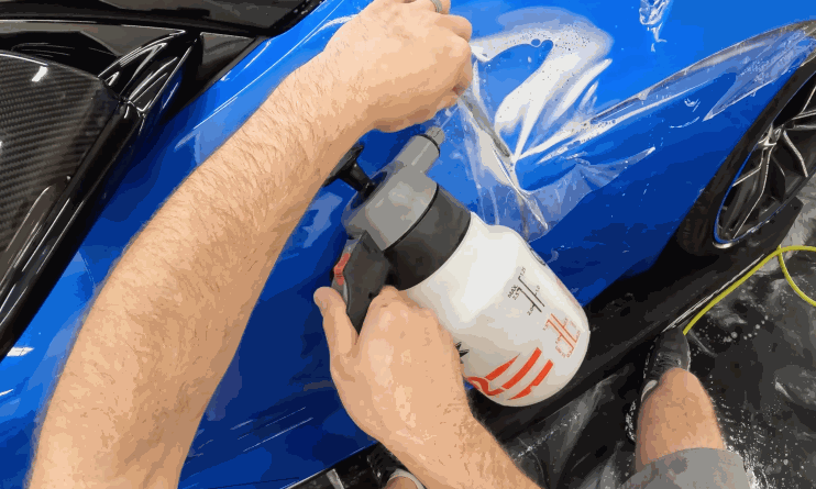 A technician sprays installation solution beneath paint protection film on the blue front fender of a sports car. The clear film is being stretched across the curved panel before smoothing.
