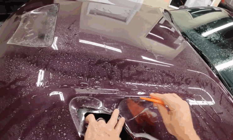 A technician uses an orange squeegee to smooth paint protection film around a recessed panel on a wet burgundy vehicle. Water droplets help position the clear film for a precise fit.