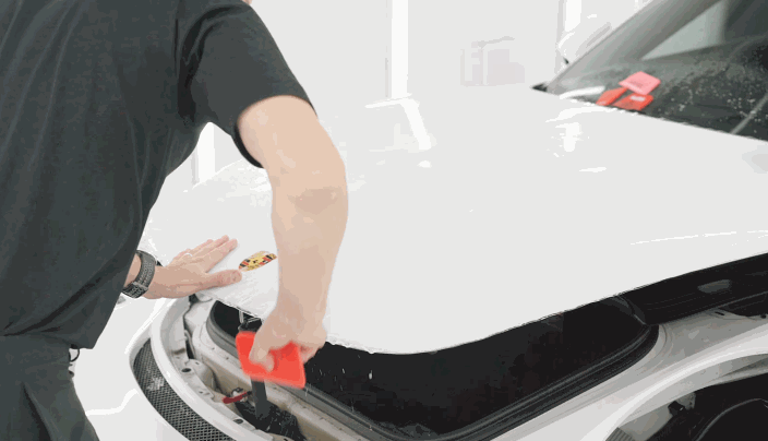 A technician smooths paint protection film across the hood of a white Porsche using a red squeegee. The clear film is fitted around the hood edges for even coverage.