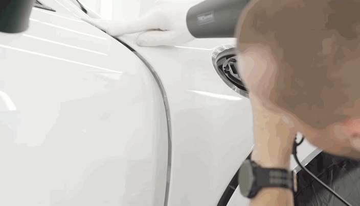 A technician uses a heat gun to shape paint protection film around the front fender and headlight of a white vehicle.
