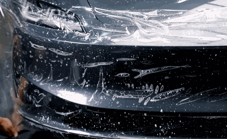 Paint protection film covers the front bumper of a dark vehicle during installation. Water and soap bubbles show the clear film being positioned across the painted surface.