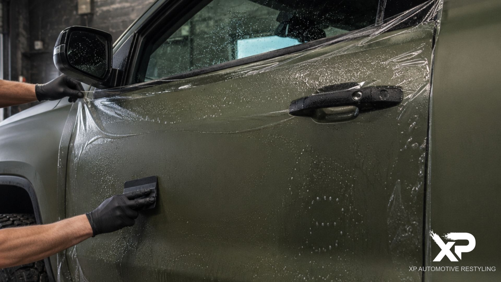 A technician applies paint protection film across the side door of an olive green vehicle using a black squeegee.