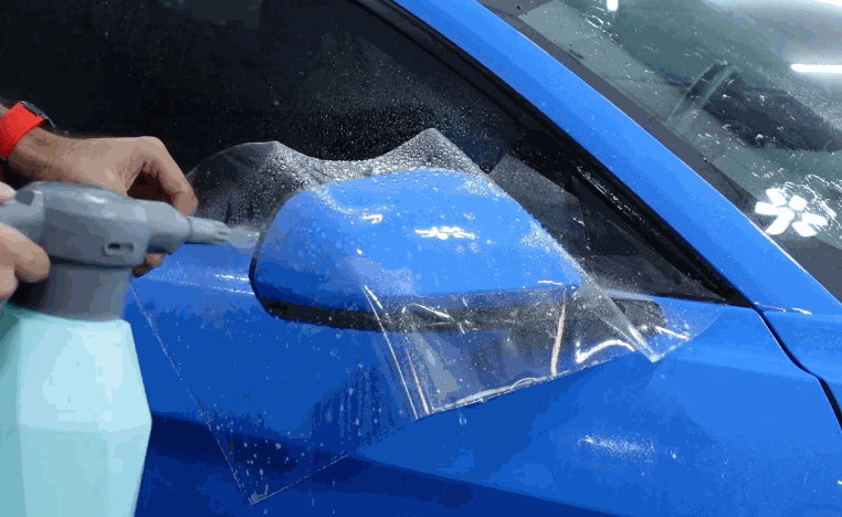 A technician sprays solution over paint protection film being fitted around a blue side mirror.