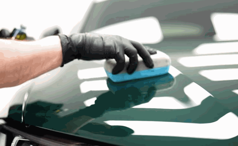 A gloved hand smooths a blue applicator across a dark green hood during ceramic coating prep.