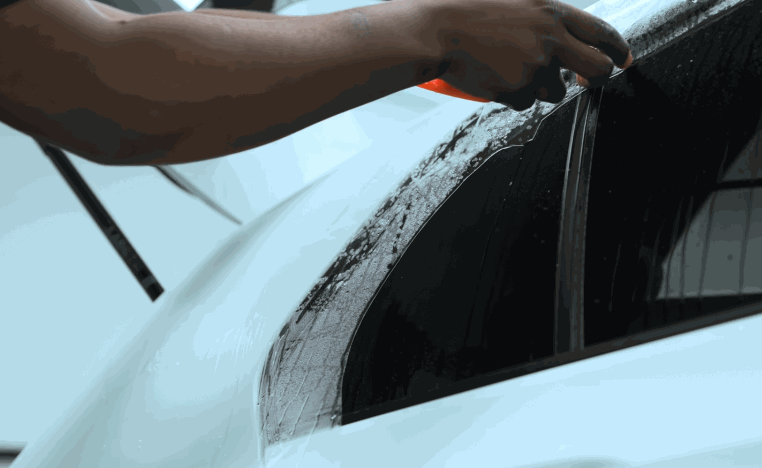 Technician applying paint protection film along the rear quarter window and black trim of a white vehicle.