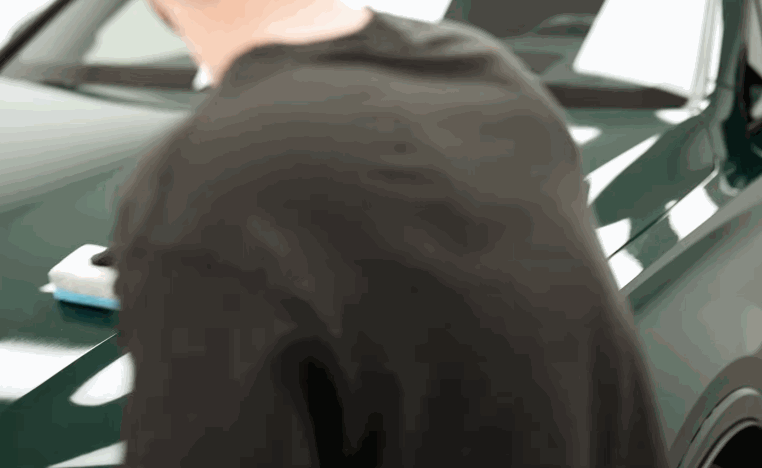 Detailer works on a glossy green car hood with an applicator pad in a detailing shop.