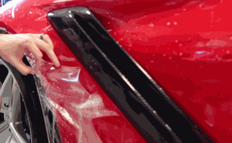 Installer smooths paint protection film over the red fender of a car near a black side vent.