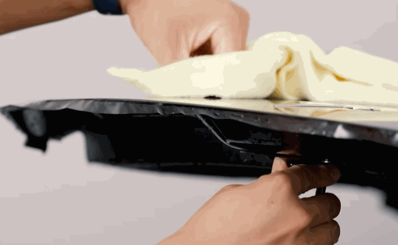 Installer trims paint protection film along the edge of a glossy black car part with a cloth resting on top.