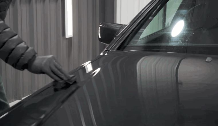 Detailer wearing a black glove wipes the gray door panel of a car under bright detailing lights while preparing the paint for ceramic coating.
