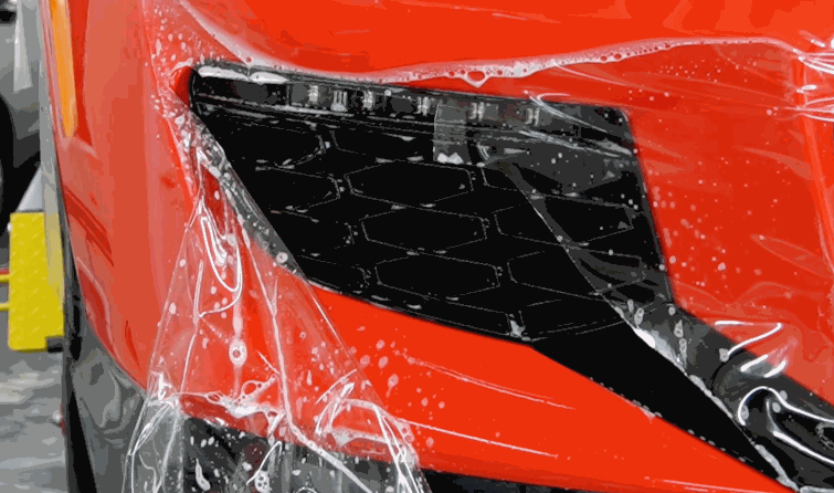 Clear paint protection film is fitted around the front vent and bumper of a red luxury car with application solution visible beneath the film. The transparent layer is shaped closely around the detailed bodywork to protect exposed paint.