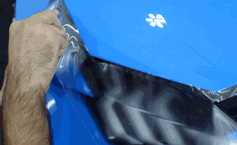 Installer carefully stretches clear paint protection film around the headlight and front corner of a blue luxury car. The film follows the contours of the bumper and painted panels for close coverage.