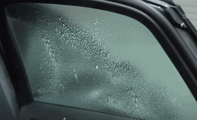 Close view of a dark car window covered with fine moisture droplets that make the glass appear hazy. The window tint remains visible beneath the condensation.