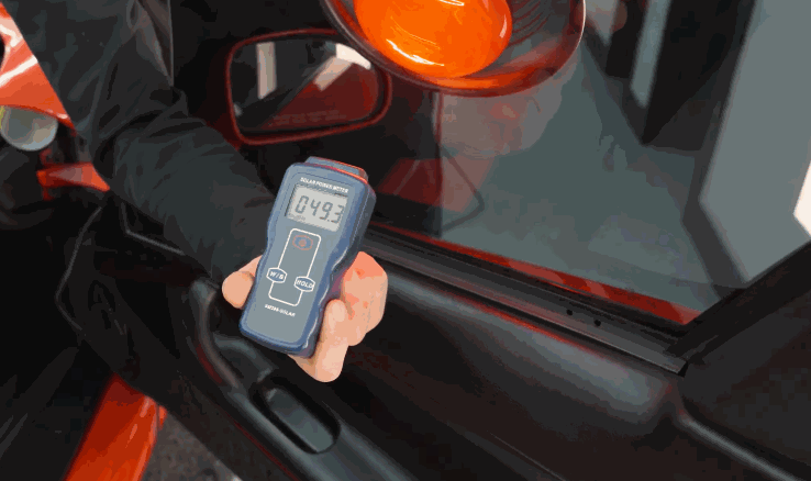 Technician holds a handheld meter against a tinted car window to measure the amount of light passing through the glass.